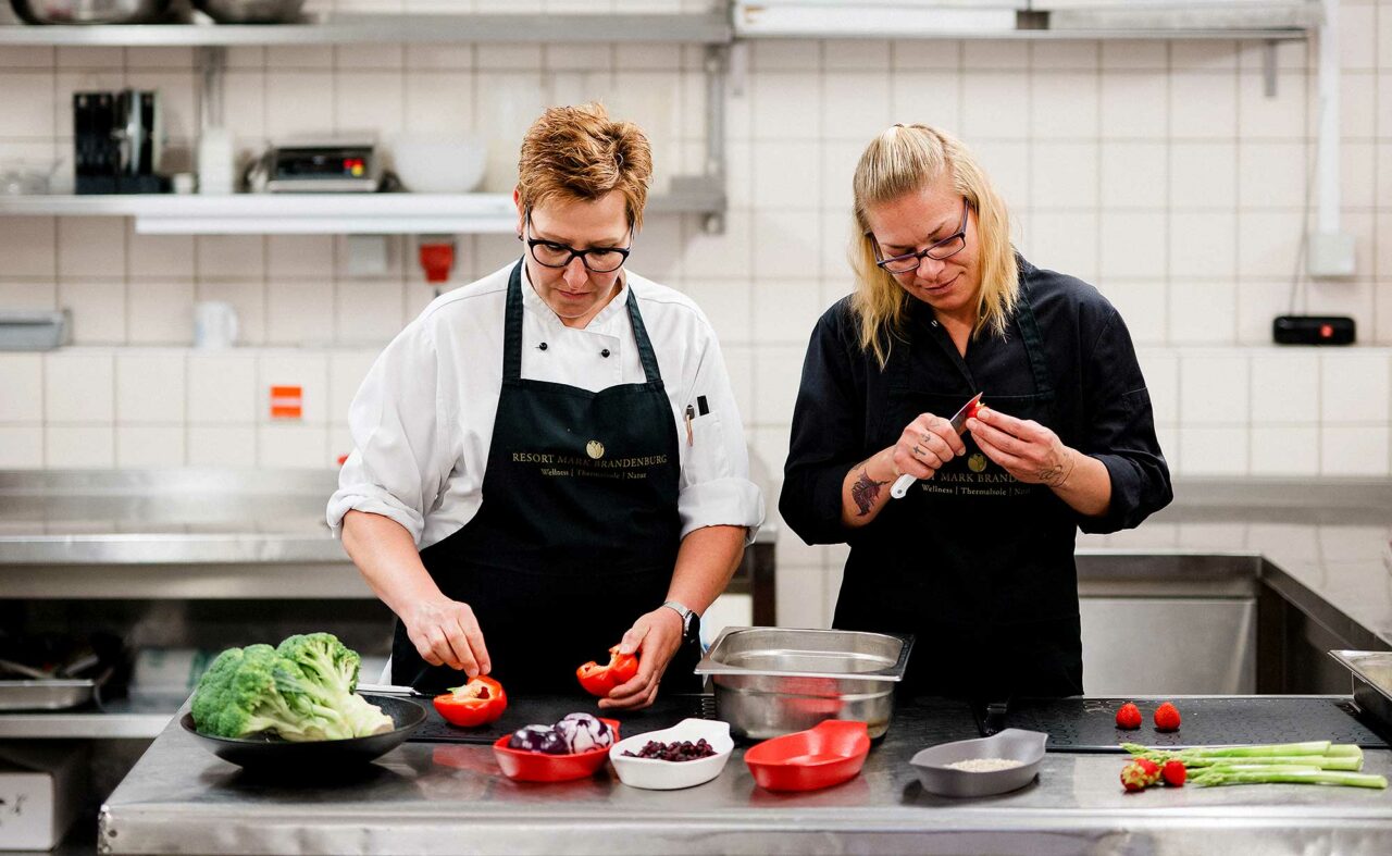 Zwei Mitarbeiterinnen vom Resort Mark Brandenburg stehen in der Küche und schneiden Gemüse klein.